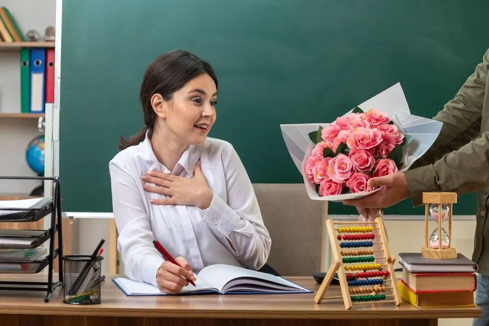 surprised-putting-hand-heart-young-female-teacher-writes-notebook-gives-bouquet-by-someone-sitting-table-with-school-tools-classroom_141793-113967.jpg
